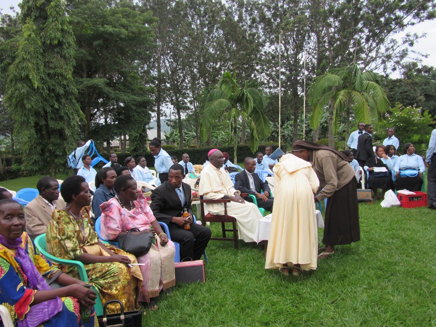 Sr. Carolyne, with her back to camera, serves Bishop Kakooza some refreshments before the concert begins. Women seated to the left are wearing the traditional gomesi.